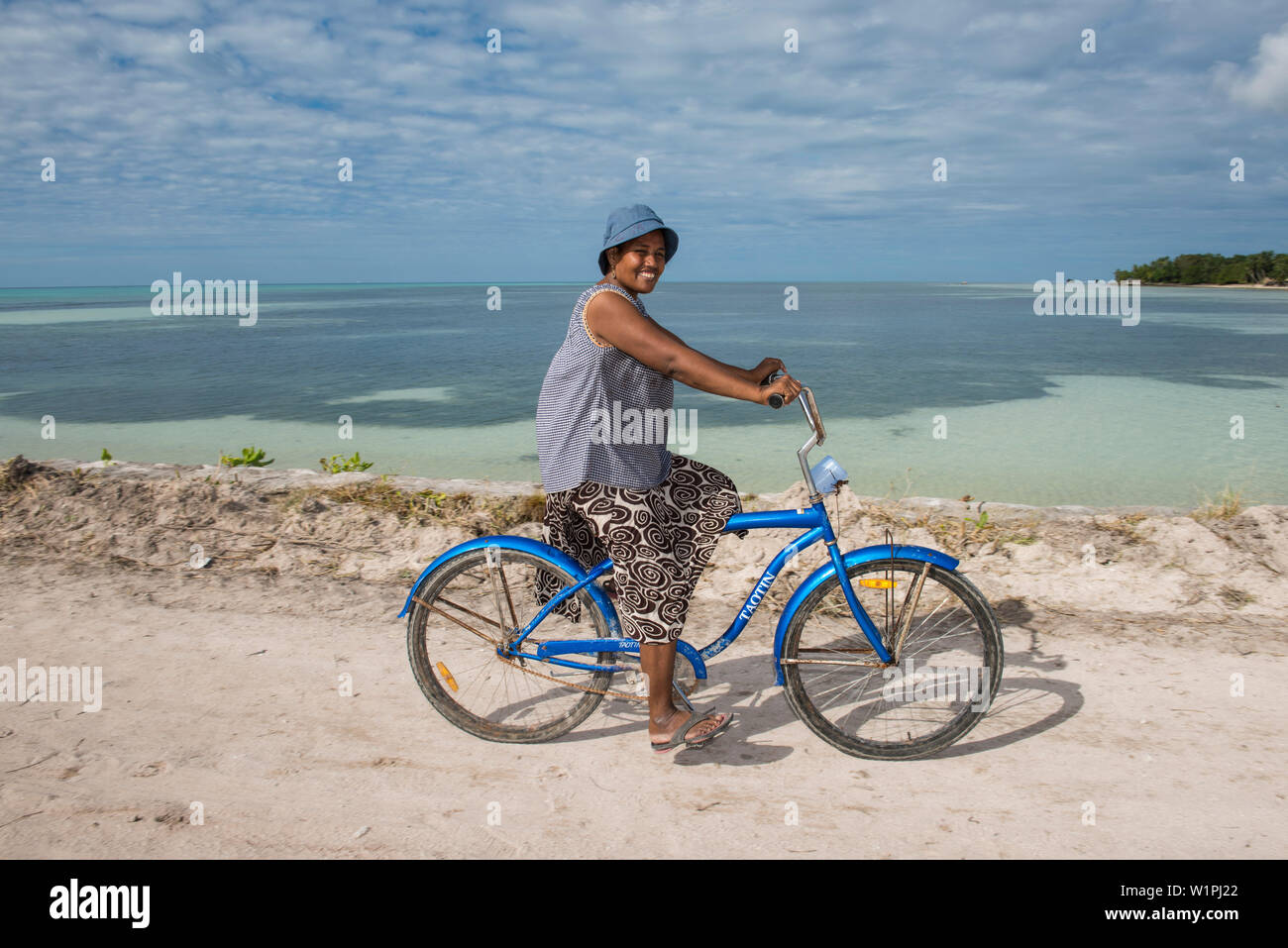 A woman in a blue hat and long skirt rides a blue bicycle along the water, Butaritari Atoll, Gilbert Islands, Kiribati, South Pacific Stock Photo