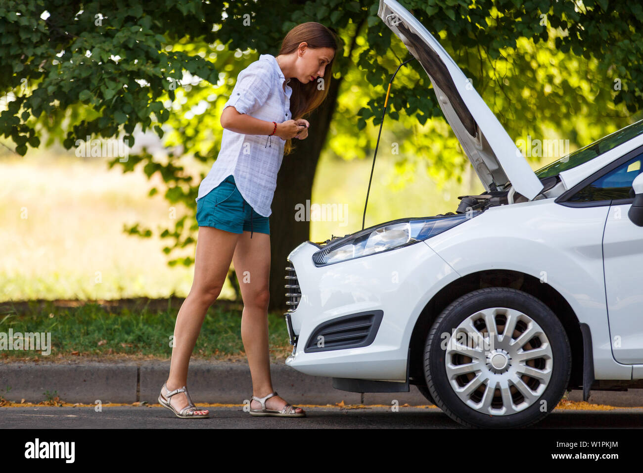 Young woman with smartphone looking at the engine of broken car Stock Photo