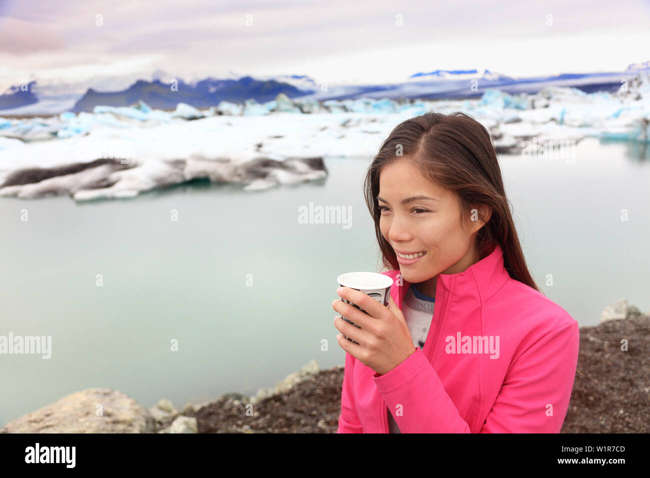 Woman drinking coffee on travel trip at glacier lagoon on Iceland. Happy tourist woman enjoying view of Jokulsarlon glacial lake. Smiling woman in beautiful Icelandic nature landscape with icebergs. Stock Photo