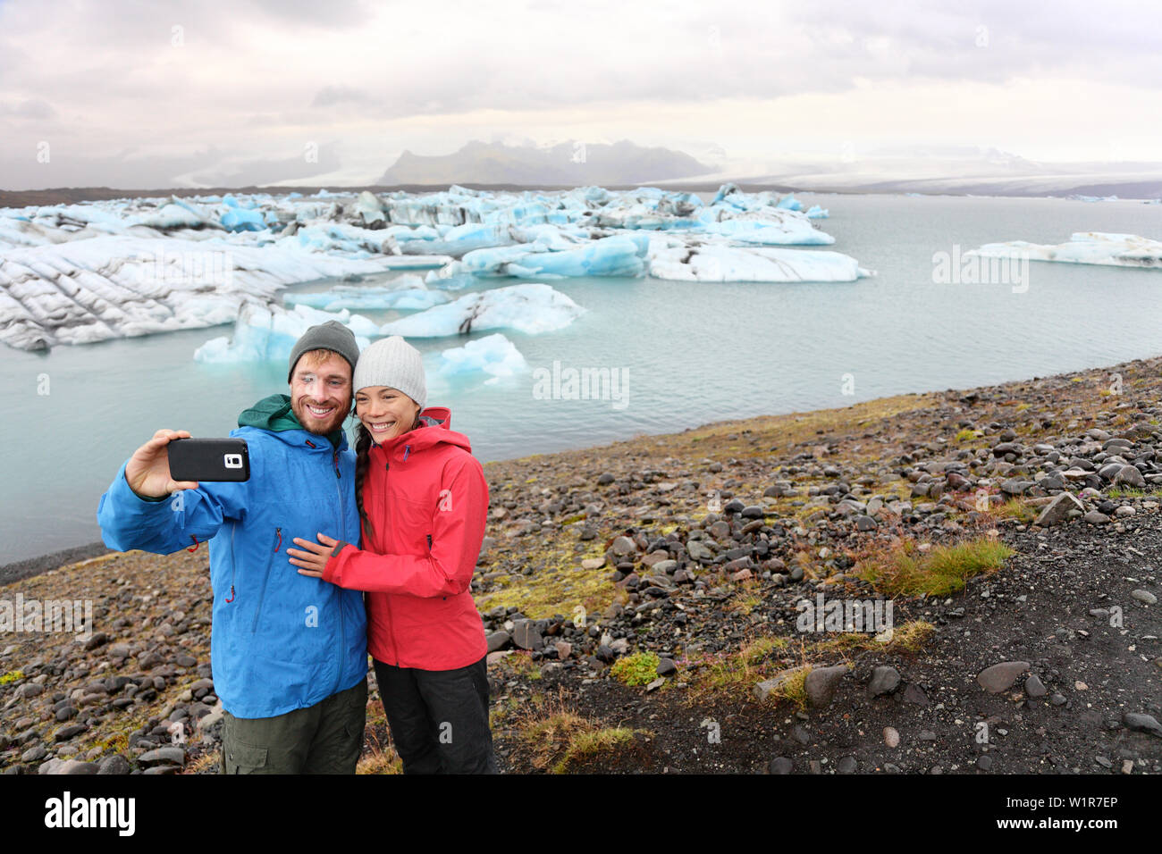 Travel couple taking selfie self portrait on Iceland photo by Jokulsarlon glacial lagoon / glacier lake. Happy tourists enjoying beautiful Icelandic nature landscape with Vatnajokull in background. Stock Photo