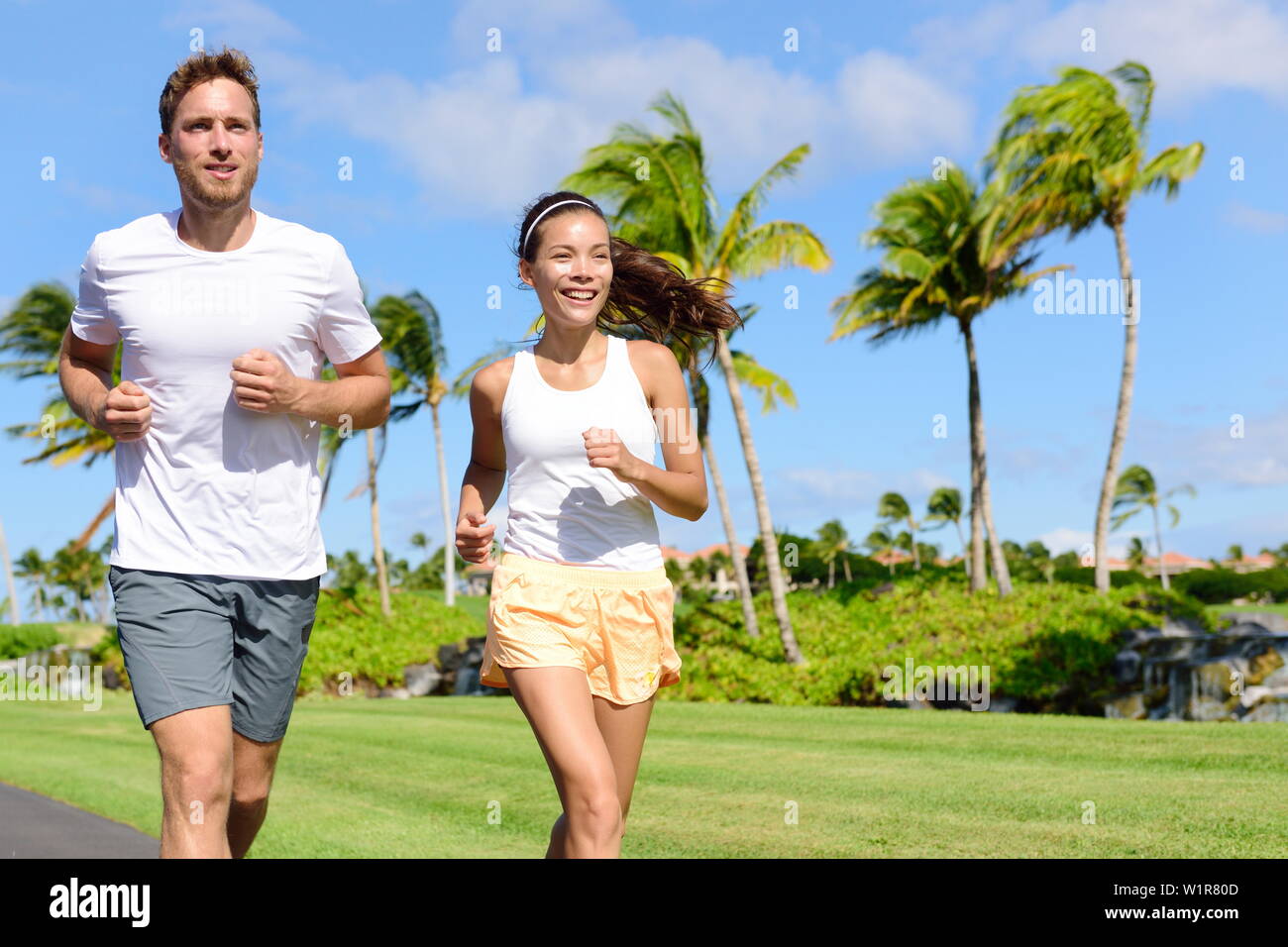 People running in city park. Happy young couple living an active healthy lifestyle  jogging training their cardio during summer on road or neighborhood street.  Multiracial group, Asian and Caucasian Stock Photo -