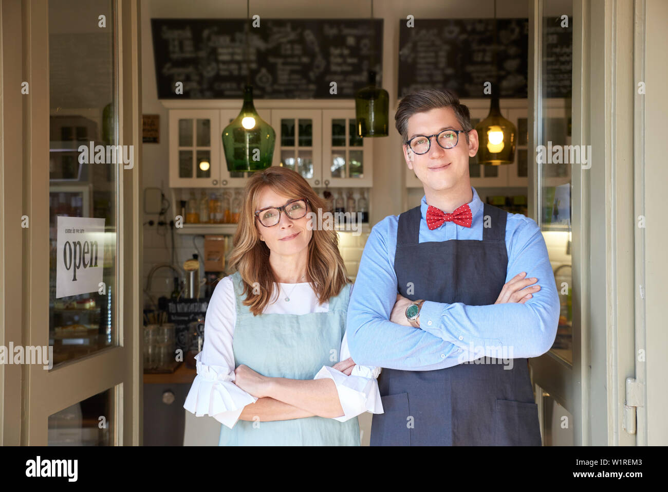 Small business owner. Shot of middle aged woman and young man wearing apron and standing with arms crossed in our small cafe’s door while waiting for Stock Photo