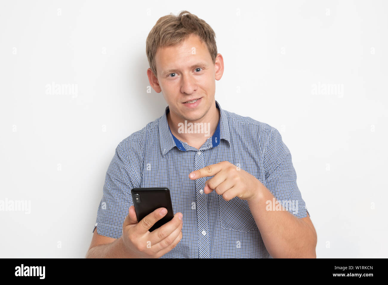 Description: Daylight Portrait of young European Caucasian isolated on white background wearing blue shirt standing in front of camera, with phone in Stock Photo