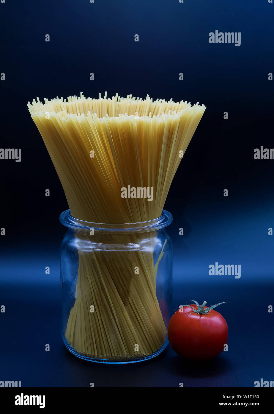 spaghetti standing in a trasparent glass jar on a blue background and a ripe tomato lying next to the can is close shot in the studio Stock Photo