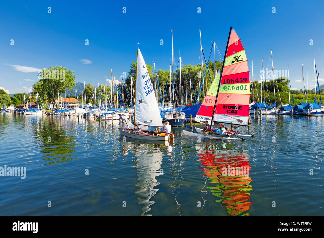Sailors in the Port field on the Chiemsee Wieser Bay; two sailing boats are reflected in the clear water; framed by blue skies and green trees Stock Photo