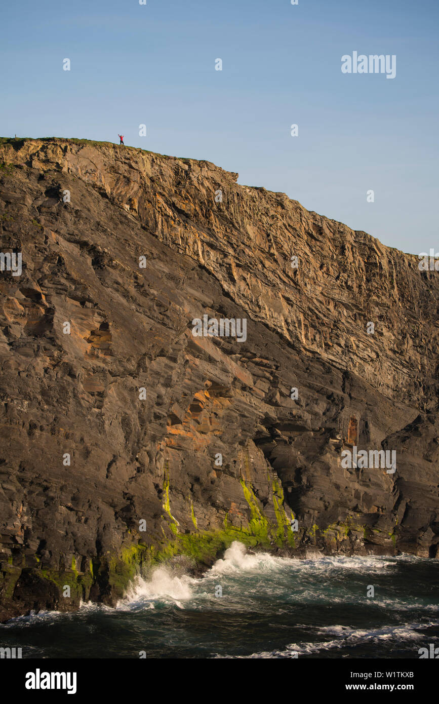 A seemingly tiny figure - a woman - stretches her arms on top of the Kilkee Cliffs and thereby puts the height in relation, Kilkee, County Clare, Irel Stock Photo