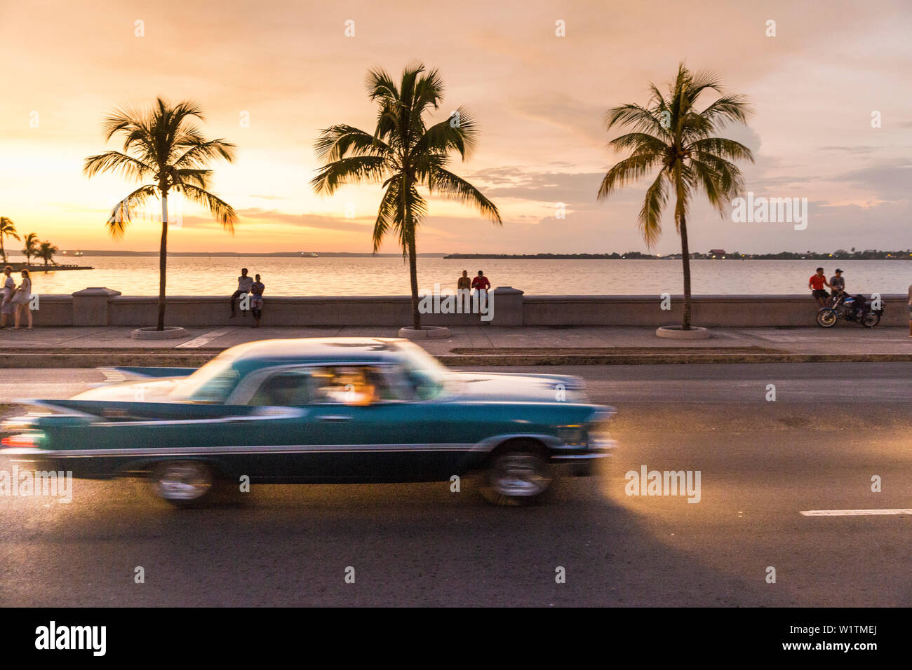 Oldtimer driving along the Malecon of Cienfuegos, meeting point in the evening and at night, nightlife, empty street, palm tree, colonial town, family Stock Photo