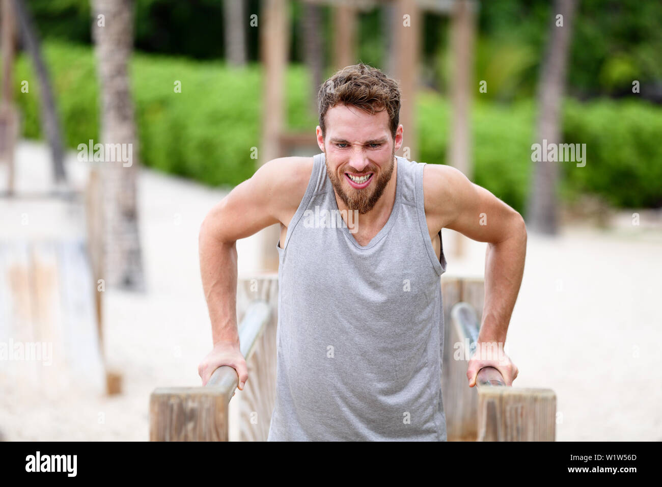 Fitness man doing dips at outdoor gym workout. Male athlete doing strength training on parallel bars in monkey jungle gym on beach. Guy exercising arms doing hard exercises. Stock Photo