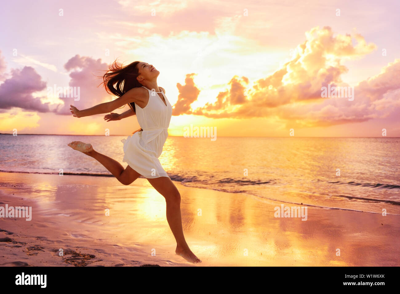 Freedom wellness well-being happiness concept. Happy carefree Asian woman feeling blissful jumping of joy on peaceful beach at sunset. Serenity, relaxation, mindfulness, stress free concepts. Stock Photo