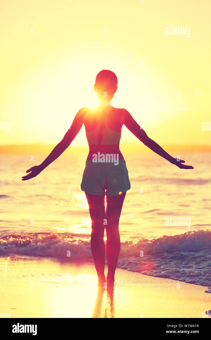 Mindfulness woman practicing yoga sun salutation at beach morning sunrise. Silhouette of fit person standing in sun flare raising arms to the sky with an open heart to do a meditation routine. Stock Photo