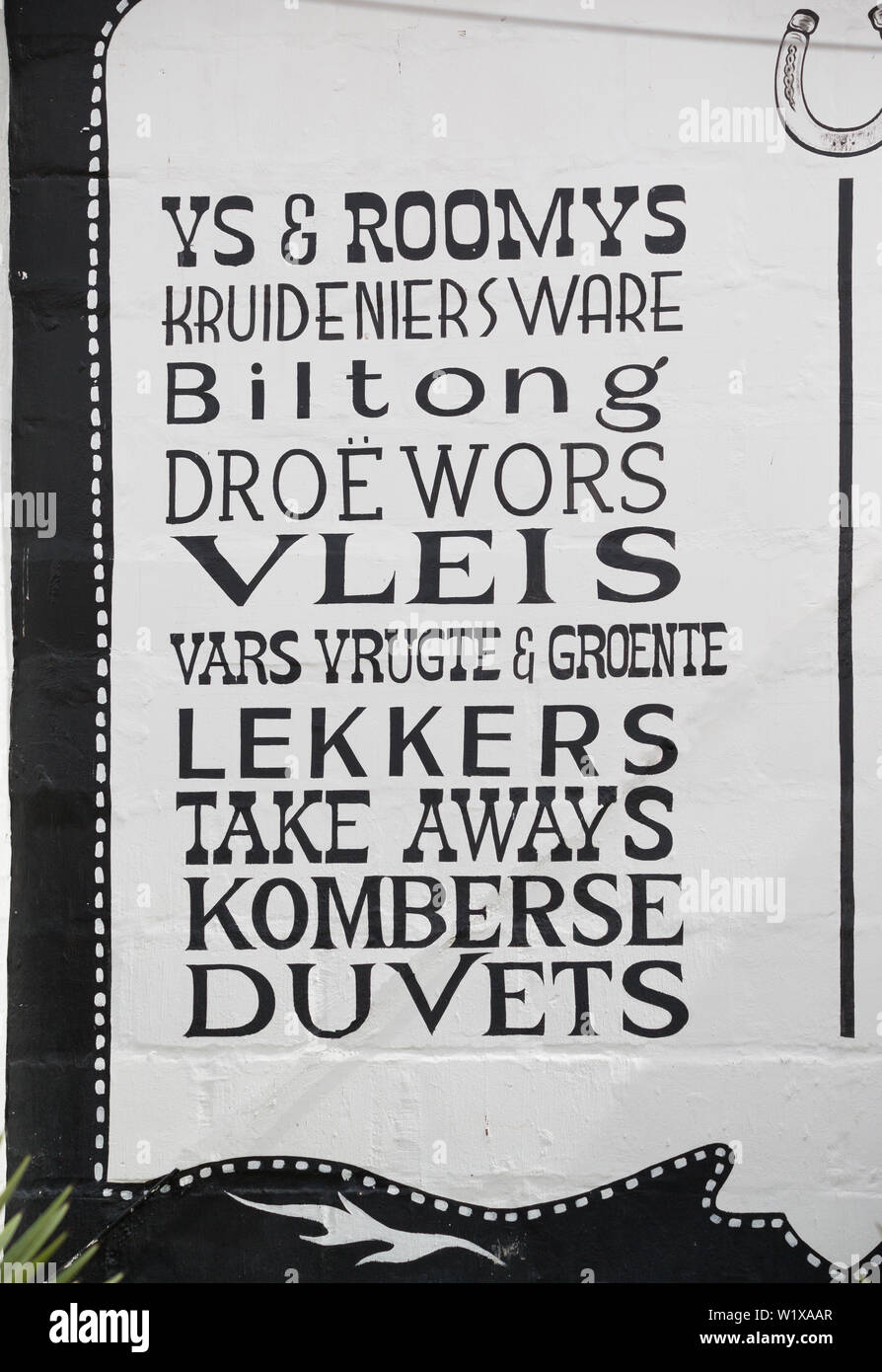 Afrikaans language words and text in a long list in black paint on a white exterior wall of a shop selling various items in South Africa Stock Photo