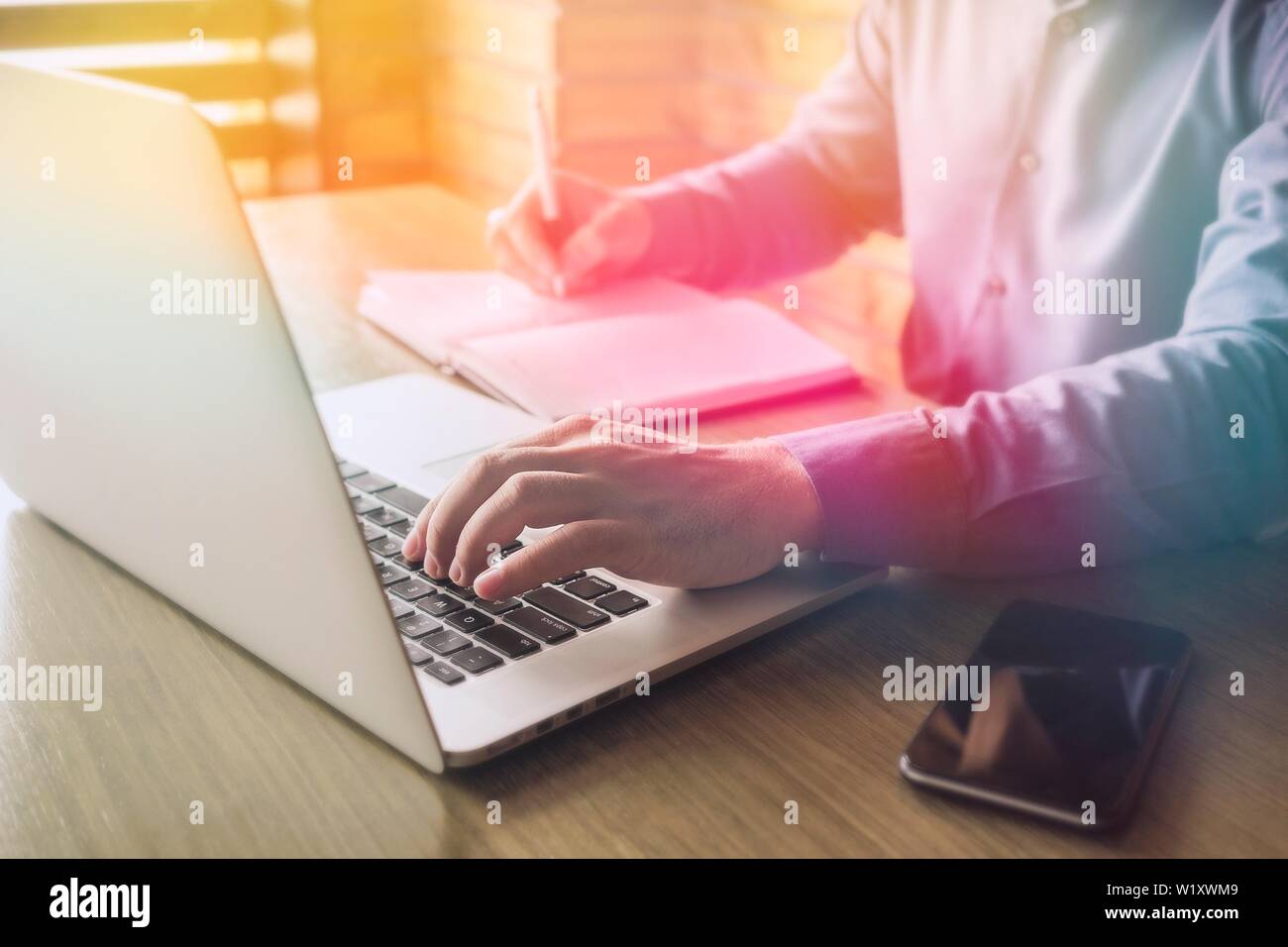 Businessman typing in information on a laptop or browsing the internet as he makes notes in a journal on the desk Stock Photo