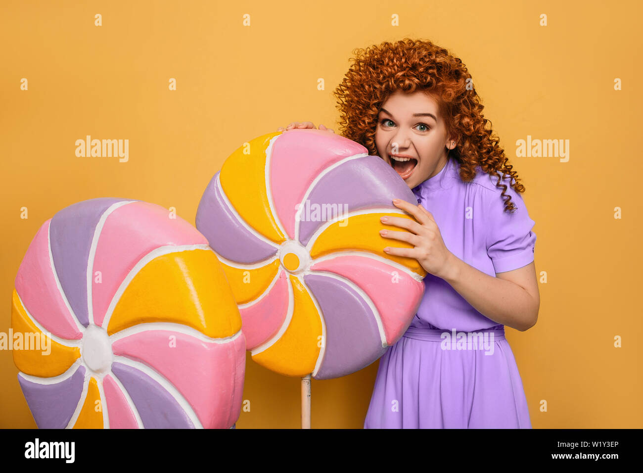 mmm yummy it is very sweet lollipop. Emotional curly haired woman bite off piece candy on yellow Stock Photo