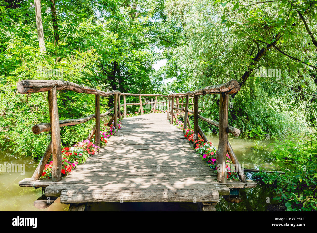 Hand made small romantic wooden bridge over the river water surrounded by trees and flowers leading to pier among river plants aged weathered wood mad Stock Photo