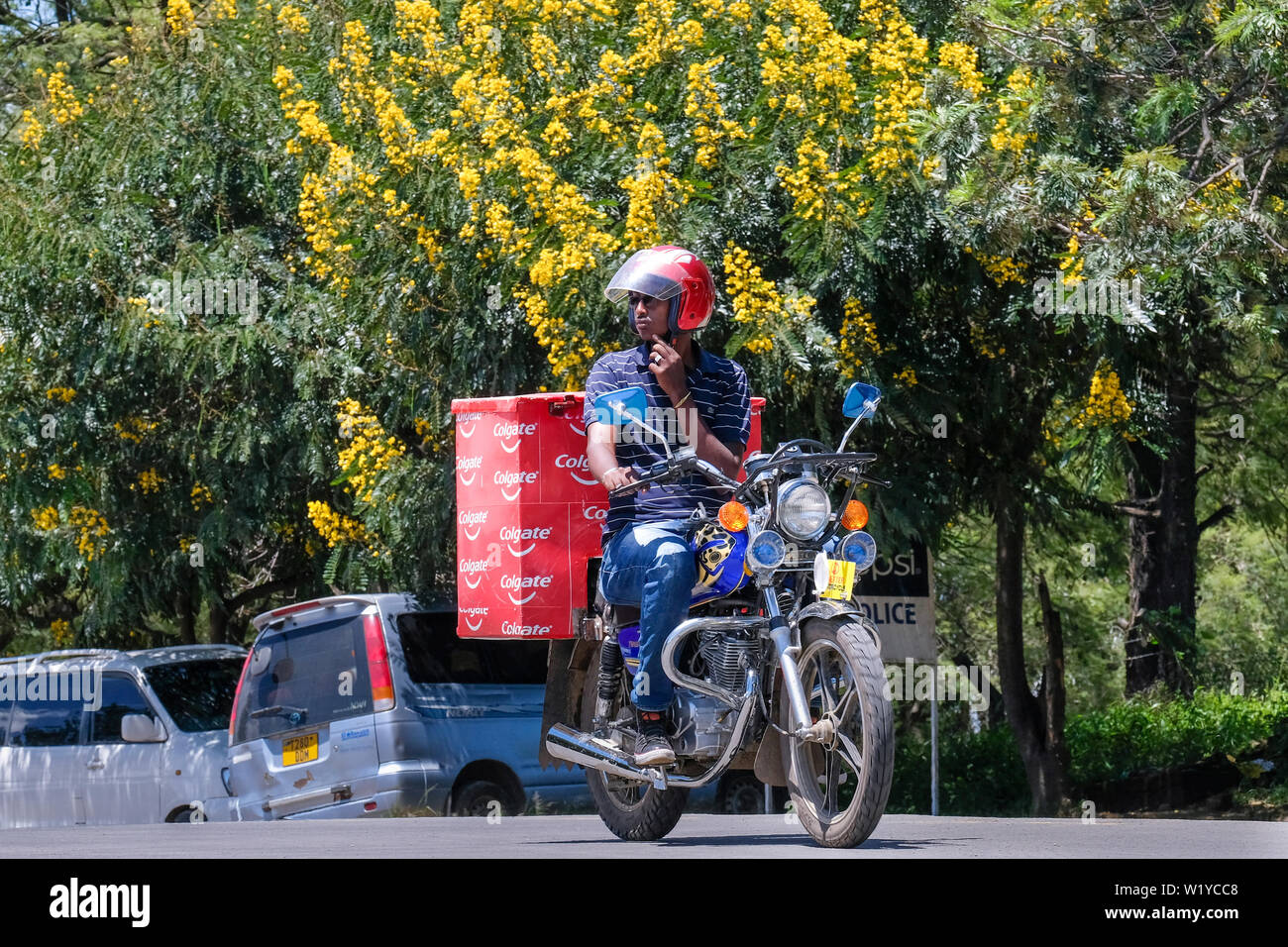 Motorcycle delivery service under yellow Jacaranda trees in the streets of Mbeya, Tanzania.   ---   Motorrad-Lieferdienst unter gelben Jacaranda-Bäumen in den Straßen von Mbeya, Tansania. Stock Photo