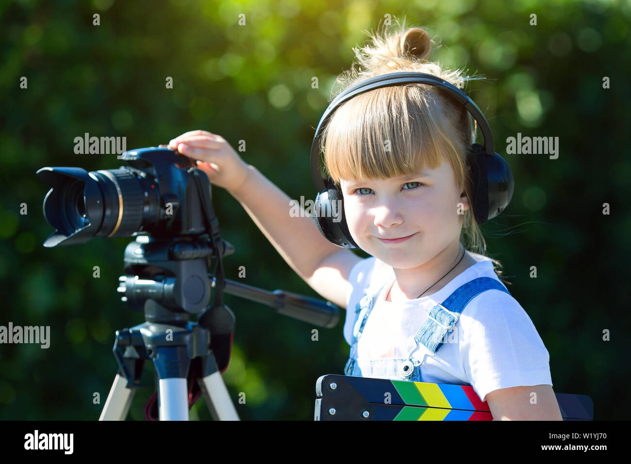 A little girl in headphones with a TV numerator in her hands is standing next camera on a tripod. Stock Photo