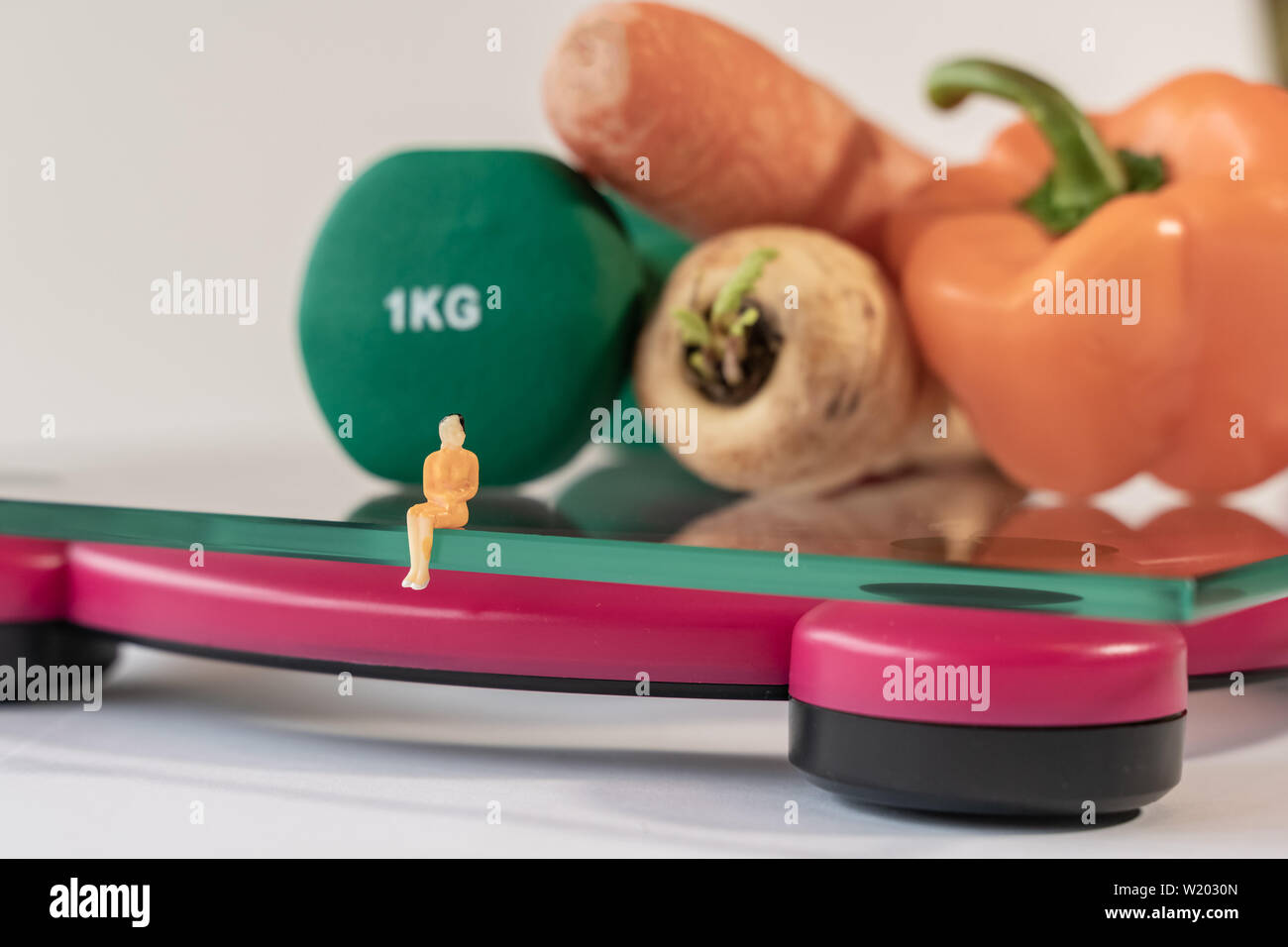 Miniature woman figure siting on the digital electronic bathroom scale for weight of human body. Fresh vegetables and green dumbbells at shallow depth Stock Photo