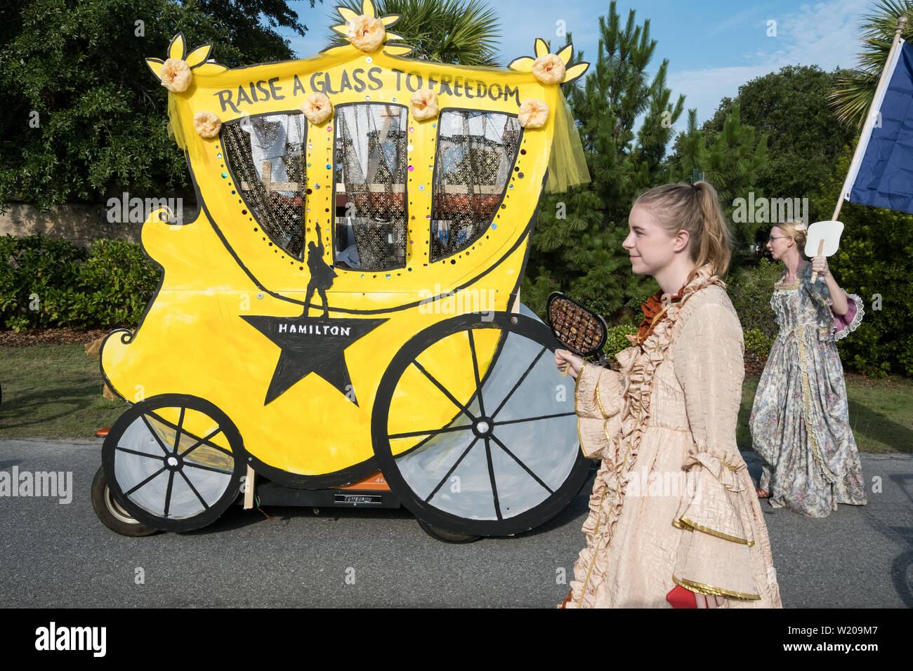 Sullivans Island South Carolina, USA. 4th July, 2019. Women wearing colonial costume walk alongside a golf cart decorated as a horse carriage during the annual Independence Day parade July 4, 2019 in Sullivan's Island, South Carolina. The tiny affluent Sea Island beach community across from Charleston holds an outsized golf cart parade featuring more than 75 decorated carts. Credit: Planetpix/Alamy Live News Stock Photo