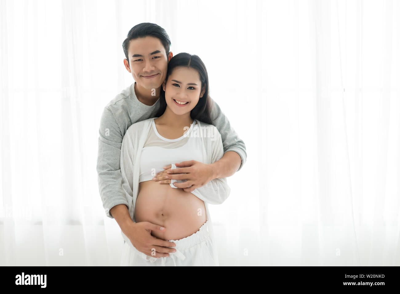 Handsome Asian man and his beautiful pregnant wife are hugging and smiling while standing near the window at home. Stock Photo
