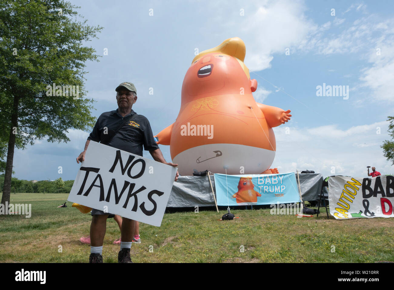 Lucio Camacho holds a sign reading “No Tanks” in front of the “Baby Trump” blimp in Washington, DC, U.S. on July 4, 2019, to protest United States President Donald J. Trump's Salute to America speech. The group believes the president's participation in 4th of July celebrations is politicizing a non-political holiday. Credit: Stefani Reynolds/CNP/MediaPunch Stock Photo