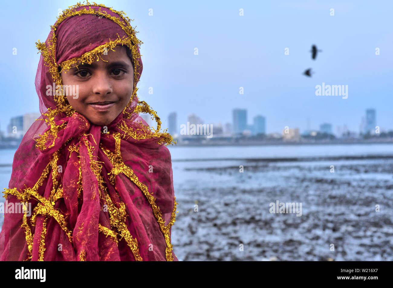 Young beggar girl on the walkway to Haji Ali Mosque, Worli Bay, Mumbai, India, on a grey cloudy day wrapped in flimsy cloth against the evening chill Stock Photo