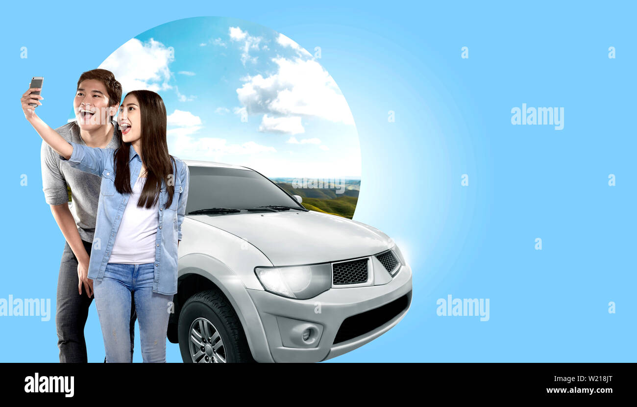 Asian couple making selfie on mobile phone camera posing beside automobile with green hills background. Traveling concept Stock Photo