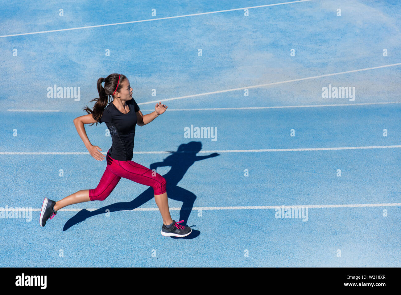 Athlete runner running on athletic track training her cardio. Jogger woman jogging at fast pace for competition race on blue lane at summer outdoor stadium wearing red capri tights and sports shoes. Stock Photo