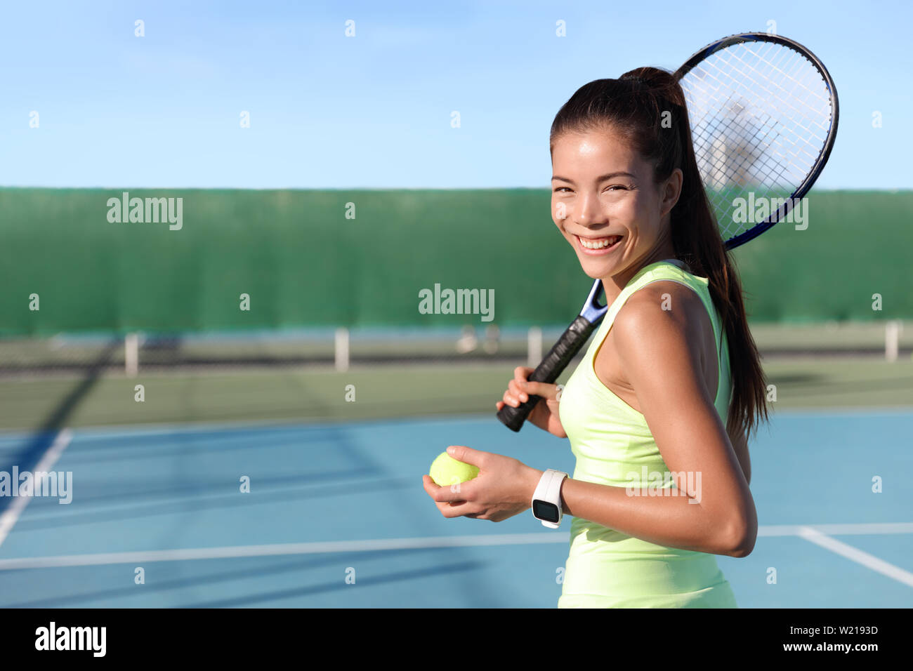 Asian young tennis player woman on outdoor hard court holding ball and racket portrait. Female athlete wearing yellow sportswear and activity tracker smartwatch. wearable tech fitness watch. Stock Photo
