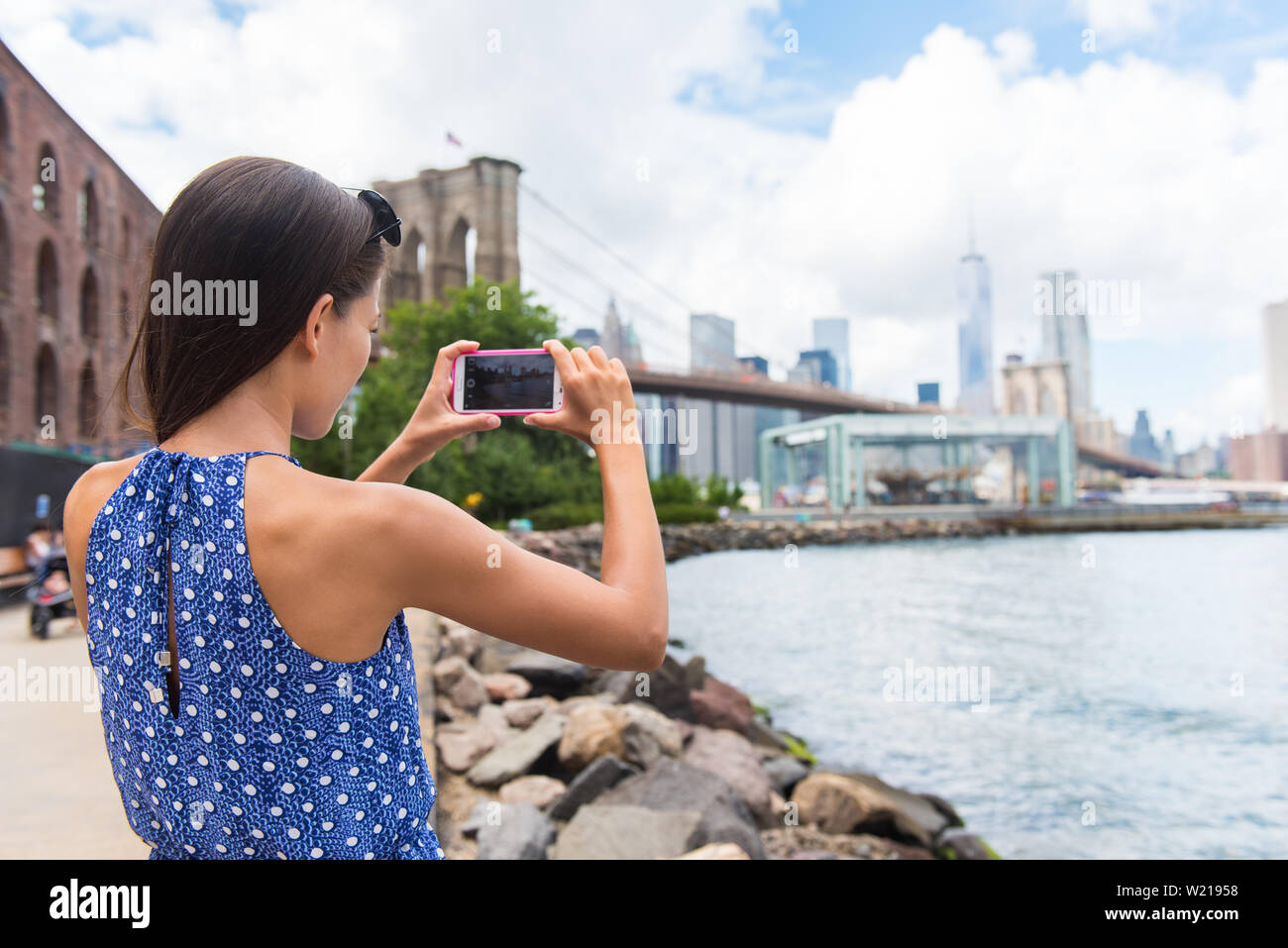 Tourist taking travel picture with phone of Brooklyn bridge and New York City skyline during summer holidays. Unrecognizable female young adult enjoying USA vacations in blue dress. Stock Photo