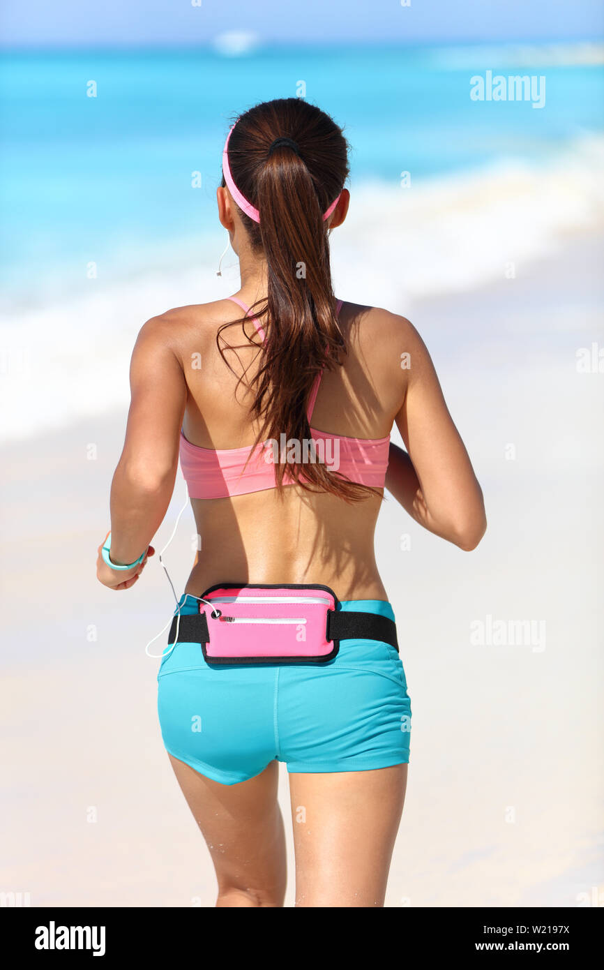 Active runner from behind jogging with wearable tech running gear fanny pack smartphone holder belt for music listening with earphones jogging away in blue sport shorts and sports bra with ponytail. Stock Photo