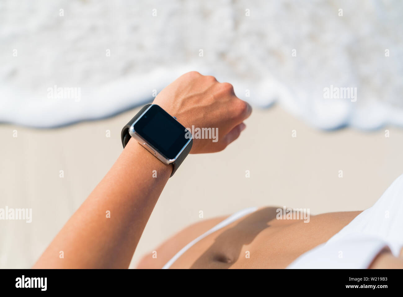 Active lifestyle woman looking at wearable tech smartwatch. Closeup of arm on beach with touch screen on wrist band of person relaxing in the water with activity tracker wrist band in outdoor living. Stock Photo