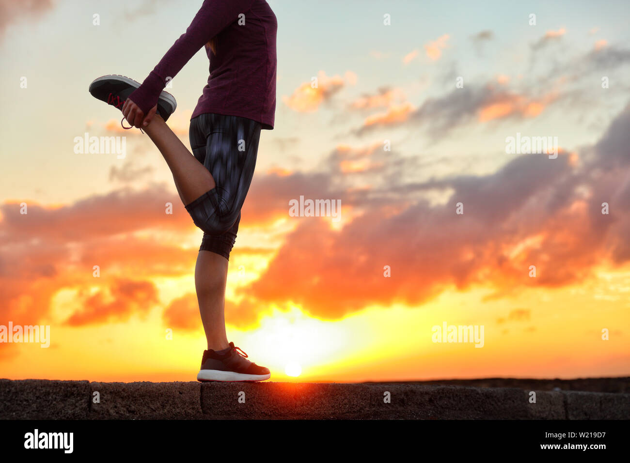 Running runner woman stretching leg muscle preparing for sunset trail run in outdoor summer nature. Female athlete lower body crop of feet doing legs stretches getting ready for cardio warmup. Stock Photo