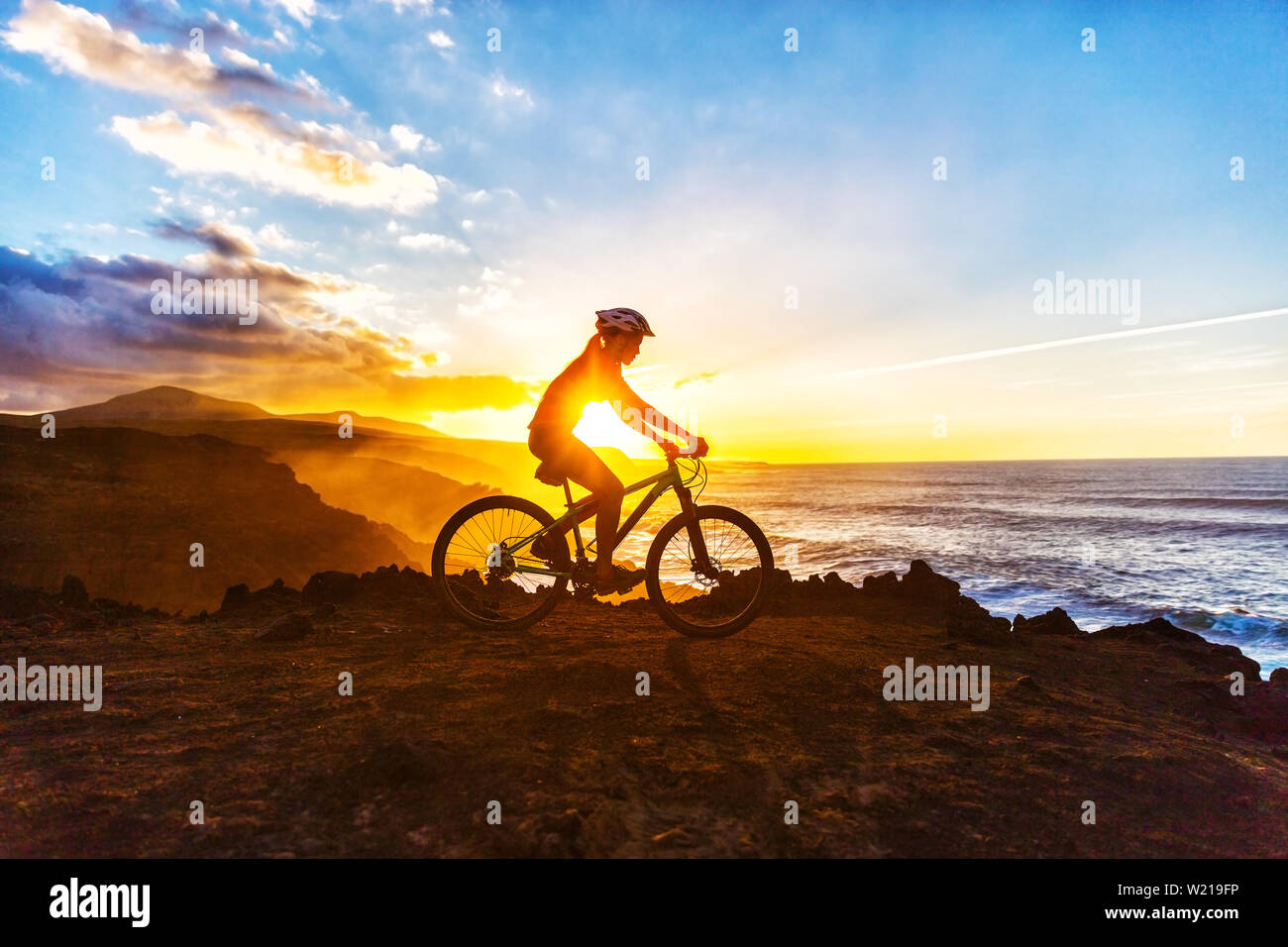Mountain biking MTB cyclist woman cycling on bike trail on coast at sunset. Person on bike by sea in sportswear with bicycle enjoying healthy active lifestyle in beautiful nature. Stock Photo