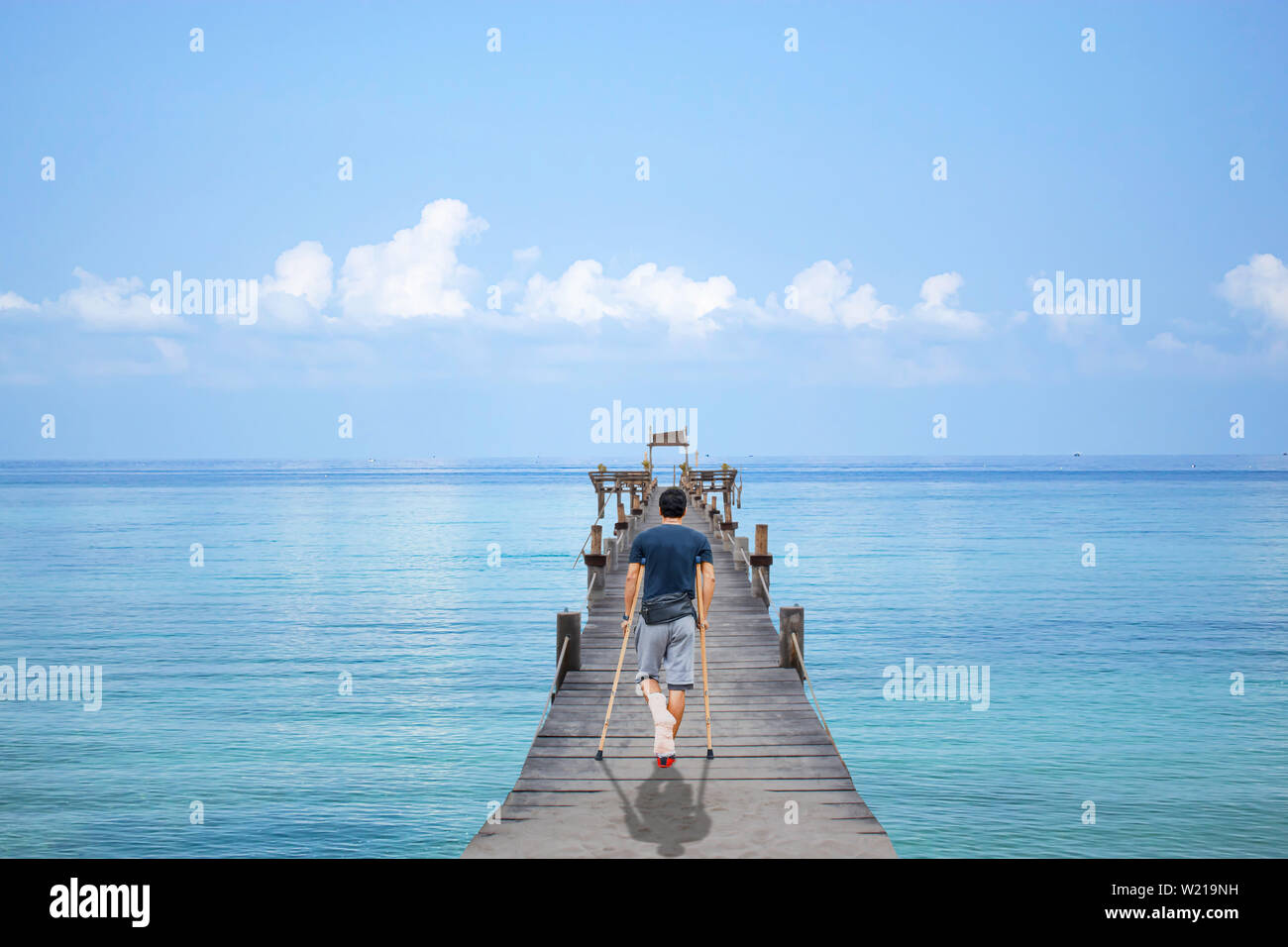 Asian man used wooden crutches walks on  bridge pier boat in the sea and the bright sky at Koh Kood, Trat in Thailand. Stock Photo