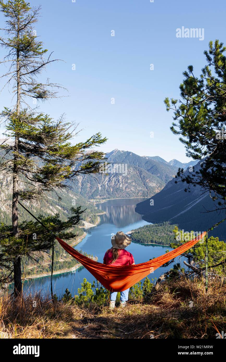 Woman with a sun hat sitting in an orange hammock, panoramic view of mountains with lake, Plansee, Tyrol, Austria Stock Photo