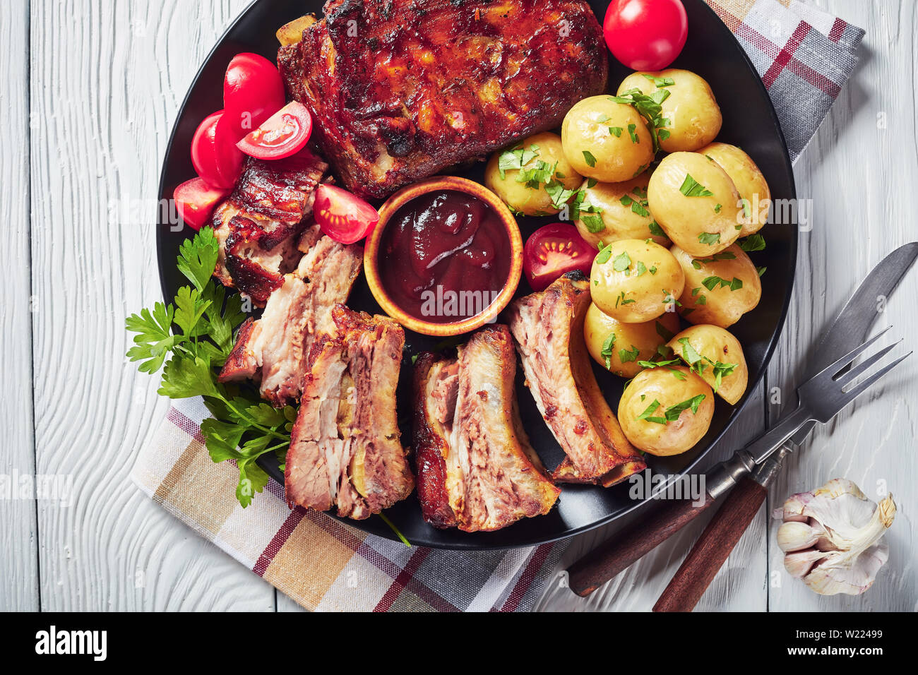 grilled pork ribs topped with barbecue sauce served with tomato salad and boiled hot new potatoes served on a black plate on a white wooden table vie Stock Photo Alamy