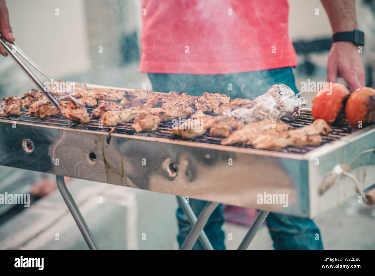 Handsome athletic man cooking barbecue outdoors. Brutal man with jeans and red shirt standing and prepared meat on a courtyard. Stock Photo