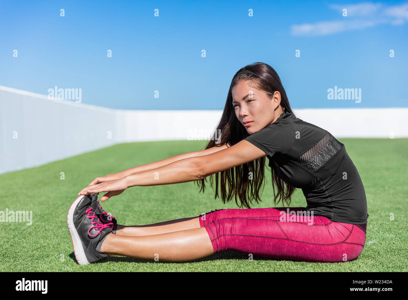 Fitness woman stretching hamstring leg muscles - back stretch sitting toe touch stretches. Seated forward bend. Sporty young athlete in activewear exercising flexibility on grass in sunny outdoor gym. Stock Photo