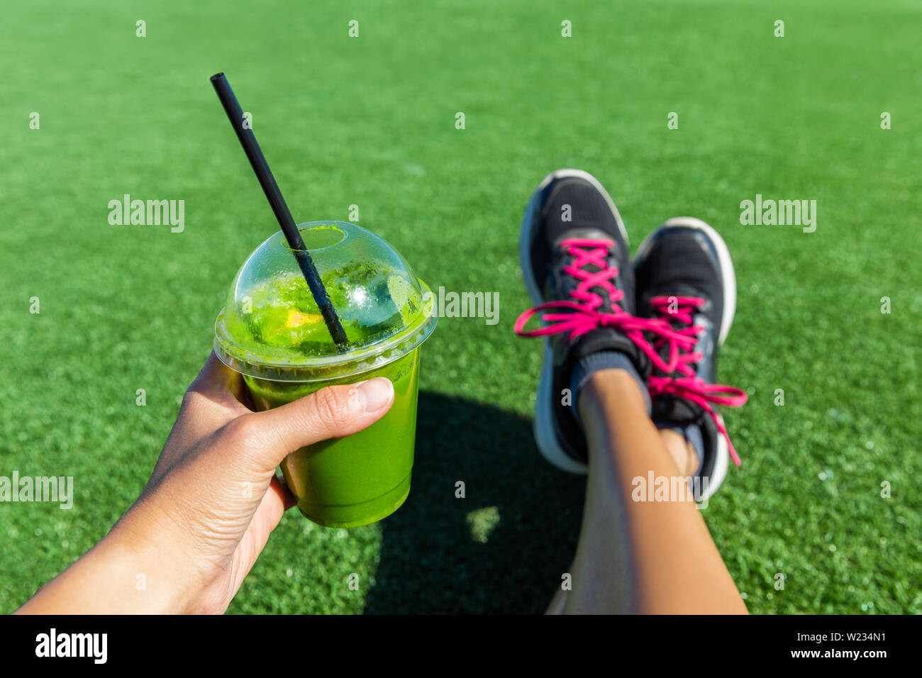 Green smoothie woman drinking plastic cup breakfast takeaway juice to go after morning run in summer park. Healthy lifestyle sporty person pov of hand holding drink with running shoes feet selfie. Stock Photo