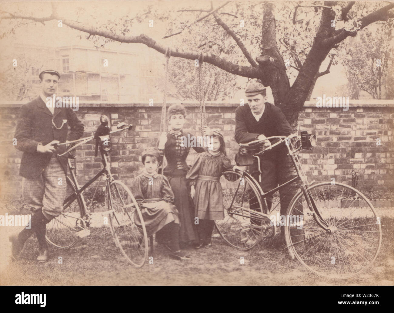 Victorian Late 1880's / Early 1890's Photograph of a Family Posing in The Garden of a House Called Pen-Isla in All Saints, Cheltenham, Gloucestershire. Lady Sat on a Tree Swing and Men Posing With Their Bicycles. This House is now No's 11 & 13 All Saints Villas Road, Cheltenham. Stock Photo