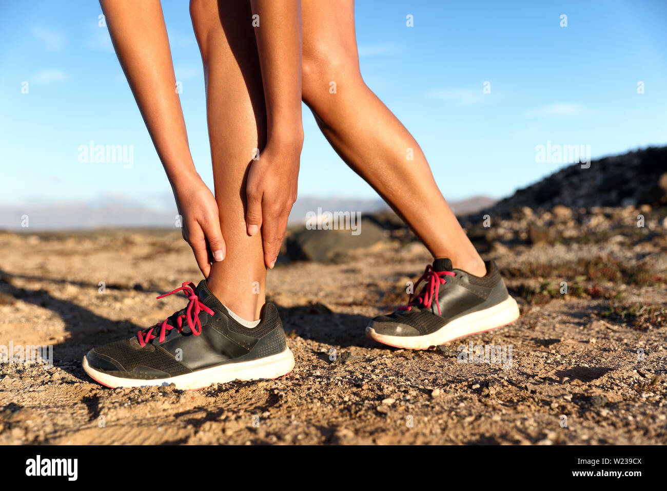 Runner woman with hurt ankles in pain during marathon. Athlete woman running outside with body injury. Sprained ankle on trail run in summer outdoors nature. Fitness leg accident on cardio workout. Stock Photo
