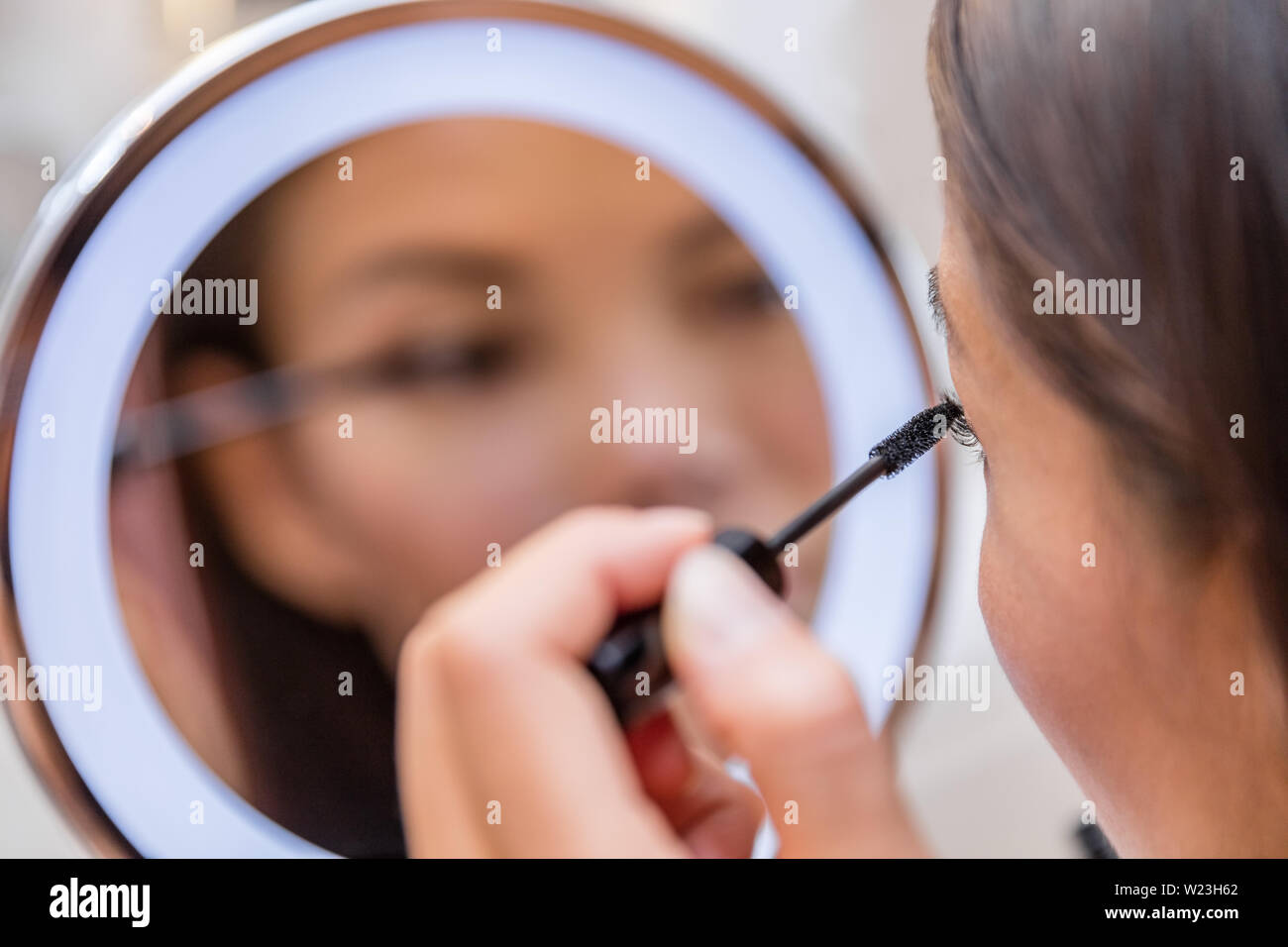 Woman putting mascara in lighted round makeup mirror from luxury hotel or home bathroom. Beautiful Asian girl getting ready for a night out applying eye make-up with brush looking at reflection. Stock Photo