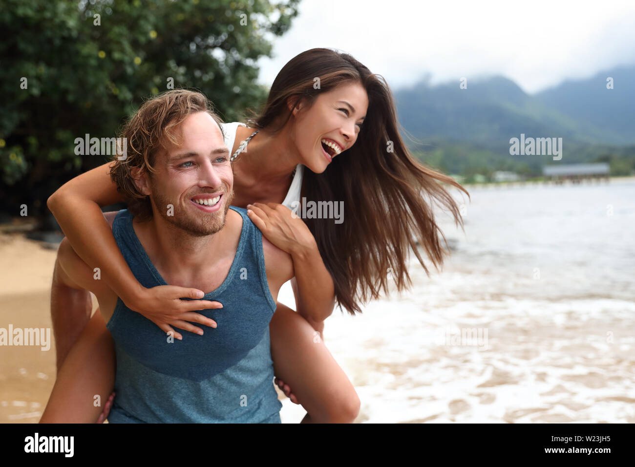 Two young people playing having fun laughing in love. Interracial couple piggybacking, boyfriend carrying Asian girlfriend doing piggyback on hawaiian beach in Kauai, Hawaii travel vacation. Stock Photo