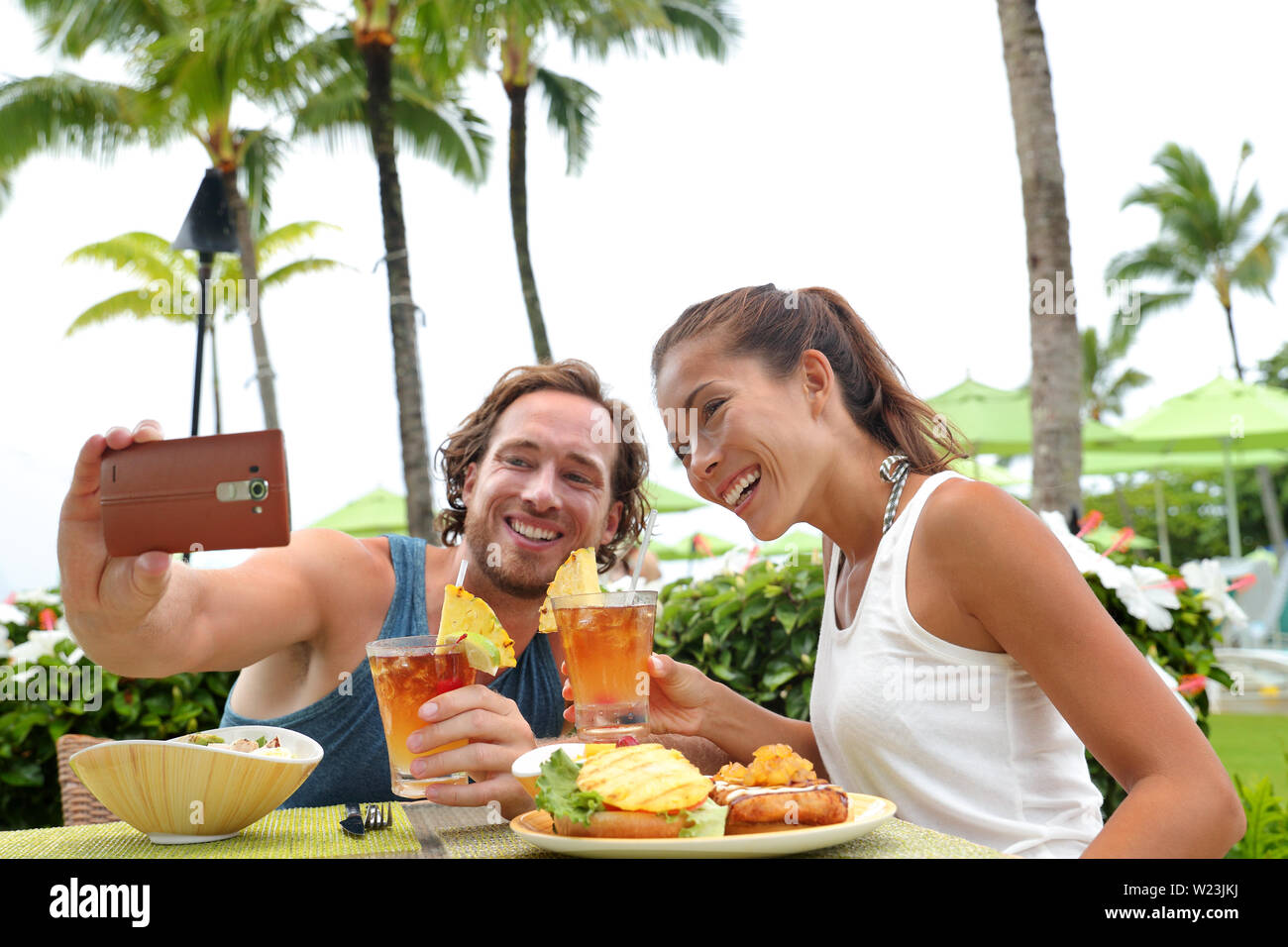 Happy young interracial couple on summer holiday going out for dinner enjoying local food meal at outdoor terrace restaurant taking a selfie picture with mobile phone for vacation memories. Stock Photo