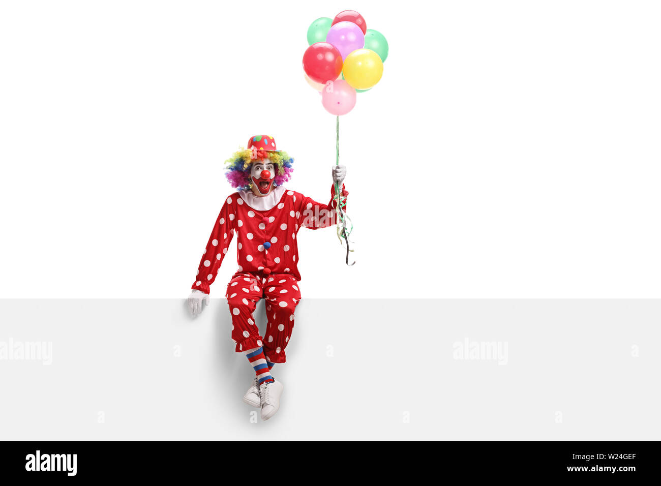 Full length shot of a cheerful clown sitting on a white banner and holding a bunch of balloons isolated on white background Stock Photo