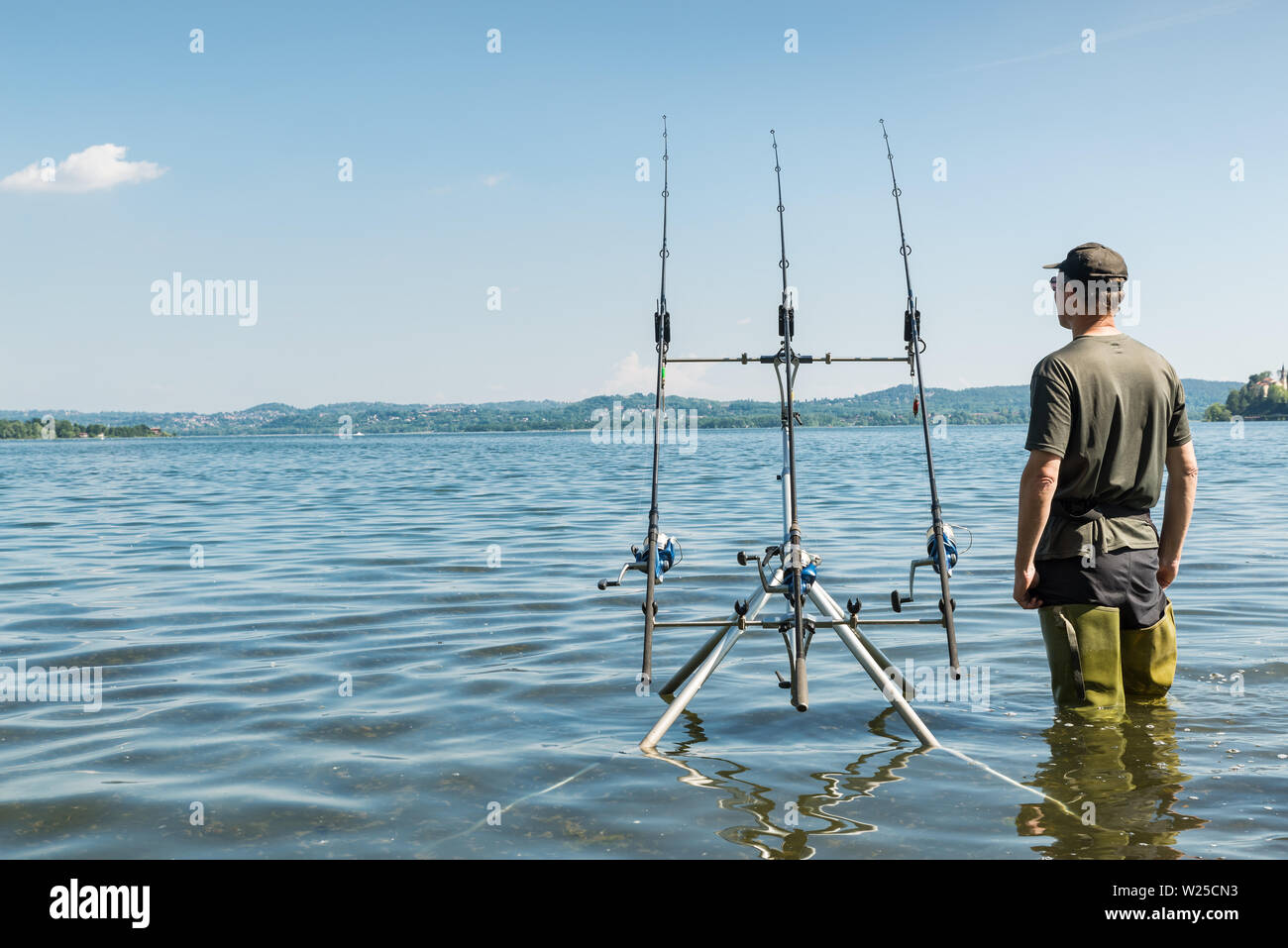 Fishing adventures, carp fishing. Fisherman with green rubber boots and camouflage clothing standing near the fishing gear. Copy space to the left Stock Photo