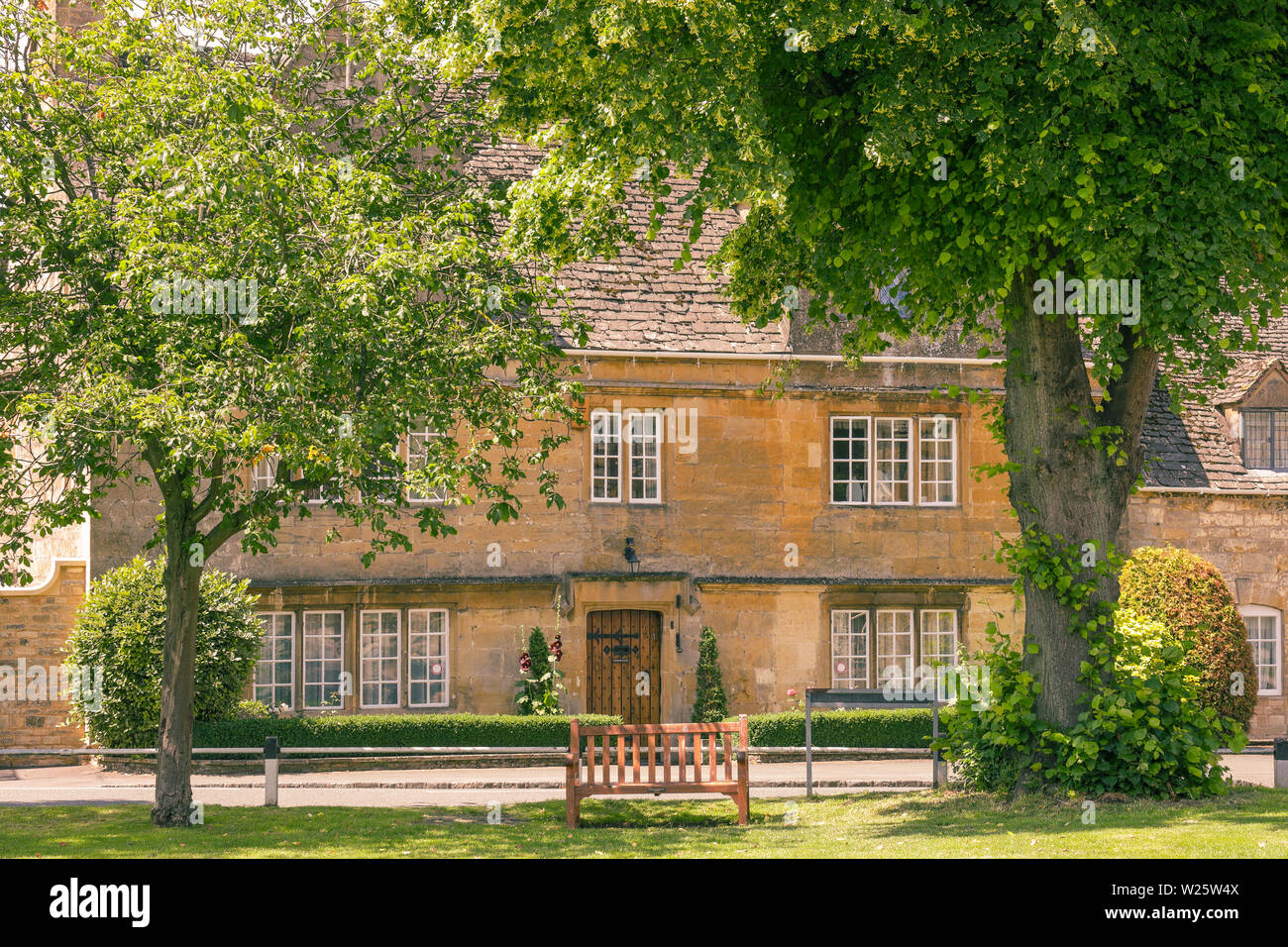 Traditional Georgian Cotswold stone cottage bathed in warm summer light facing the village green seating area with a bench Stock Photo