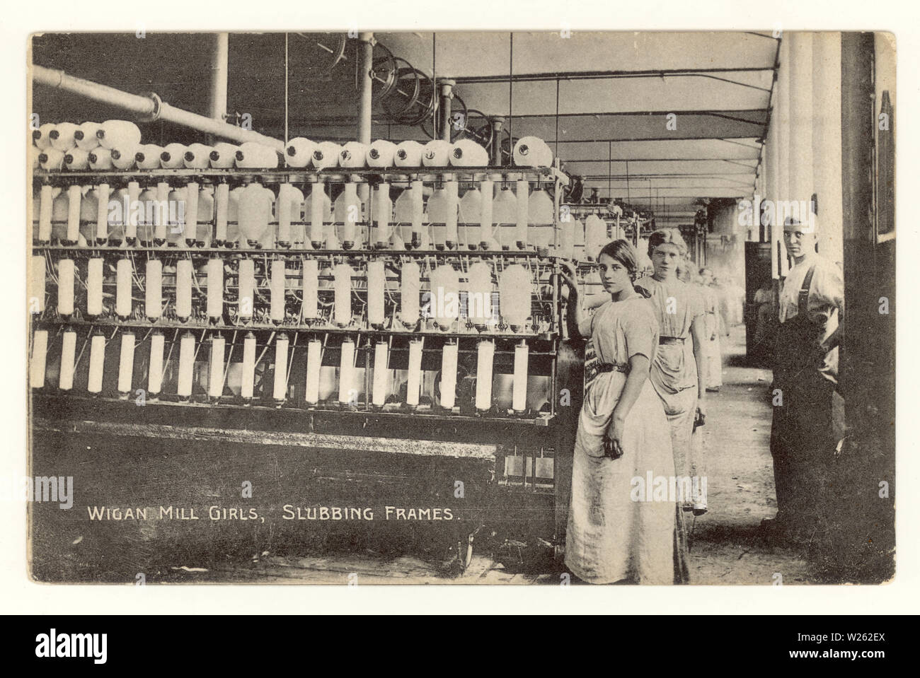 Early postcard printed at bottom is  'Wigan Mill girls,slubbing frames', with overseer / foreman standing beside slubbing frames - a machine used to straighten out the fibres in cotton prior to spinning 'Lancashire Lasses',  circa 1910, Wigan, Greater Manchester, U.K. Stock Photo