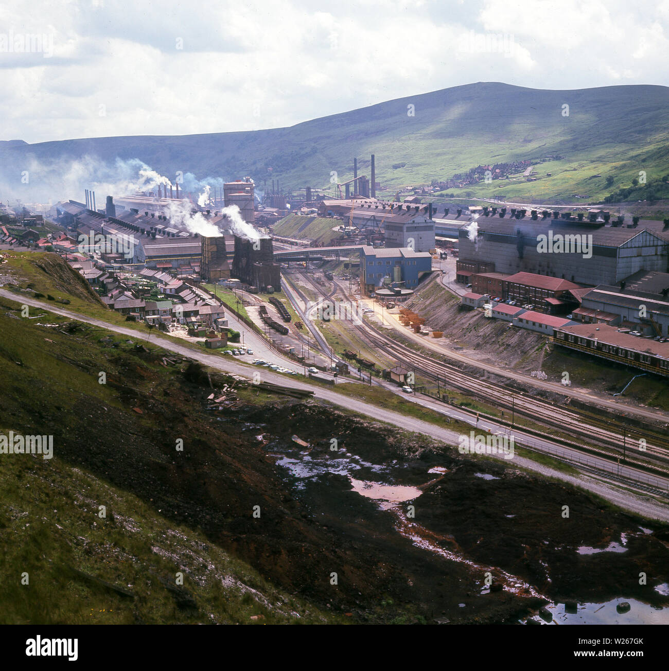 1960s, historical, view over the Ebbw Vale steelworks, Wales, showing the giant industrial complex, railway lines, workers housing and surrounding landscape, which provided both iron ore and coal. In the 1940s, the steel mill was the largest in Europe. In 1951, RTB, the company that owned the mill was nationalised. In the 1970s, due to changes in the industry and its location, remote from vast mine pits and bulk shipping facilities, steel making at the site ceased. Stock Photo