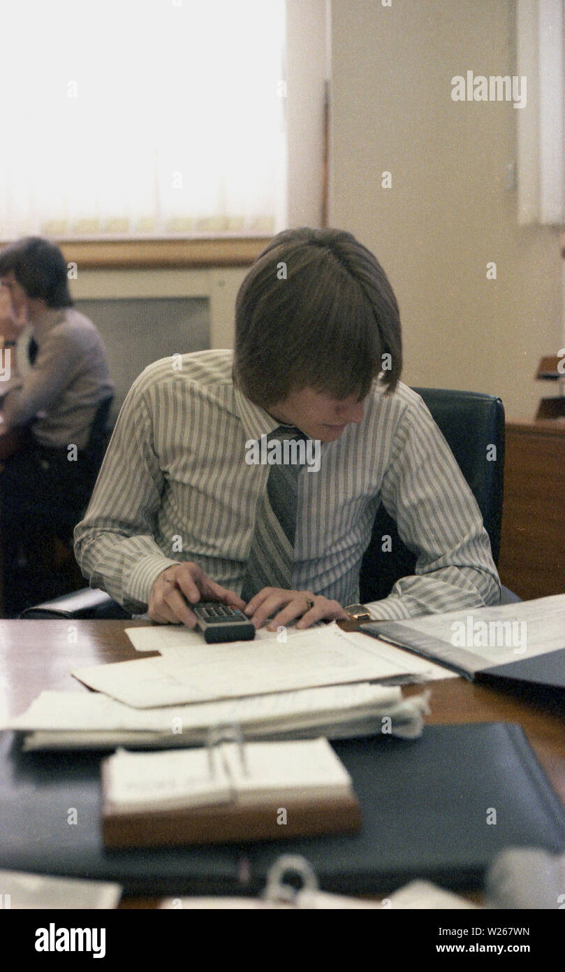 1970s, historical, a young adult male, late twenties, or early thirties in age in an office, sitting at a desk, working, a financial manager or possibly an accountant as he is using a small calculator to add figures from printed ledgers or journals, England, UK. Stock Photo